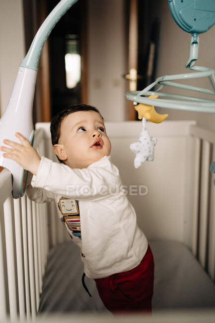 Сверху очаровательный маленький мальчик, стоящий в удобной кроватке и играющий с белой резиновой туфлей — стоковое фото