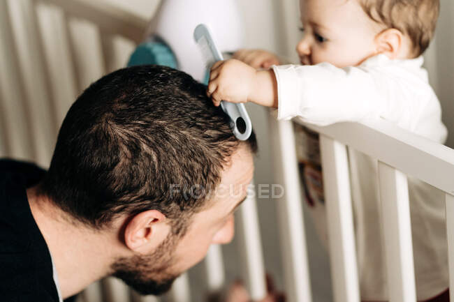Contento pequeño hijo de pie en cuna y cepillar el pelo de los padres jóvenes con peine - foto de stock