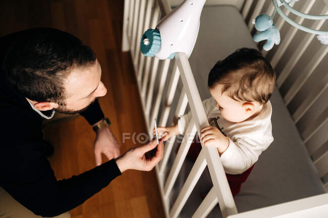 De arriba contenido pequeño hijo de pie en cuna con peine y padre joven - foto de stock