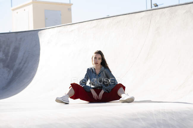 Full body young female in stylish outfit sitting in skate park while looking at camera in sunlight — Photo de stock