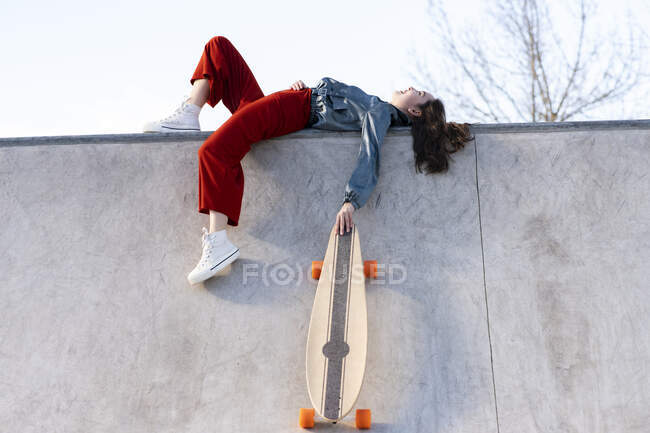 Low angle side view of tired female in trendy outfit with longboard lying in concrete skate park on sunny day — Stock Photo