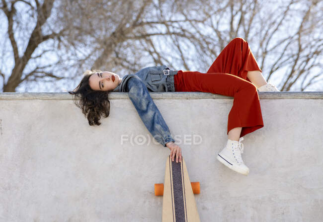 Low angle side view of tired female in trendy outfit with longboard lying in concrete skate park on sunny day looking at camera — Fotografia de Stock
