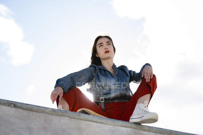 Низький кут жіночого підлітка в модному одязі і камерах, що дивиться далеко від бетонного паркану в спині, освітленому блакитним прозорим небом — стокове фото