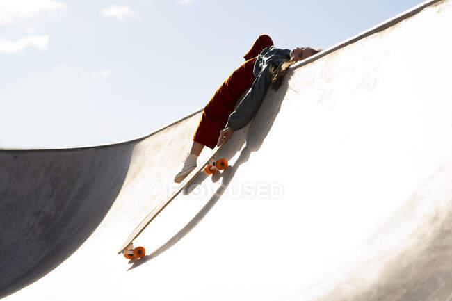 Vista lateral de ángulo bajo de mujer cansada en traje de moda con longboard acostado en el parque de patines de hormigón en el día soleado - foto de stock