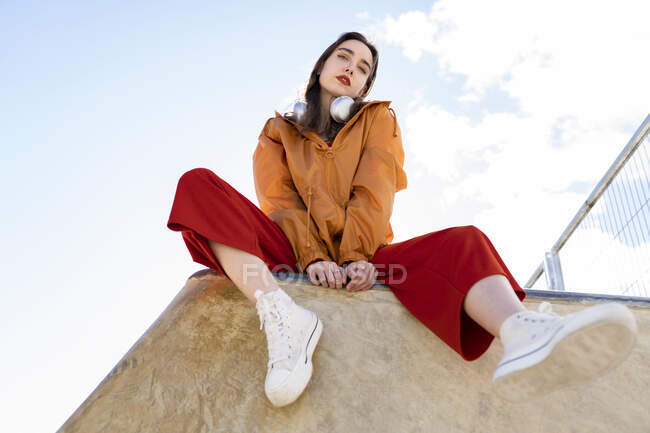Низький кут жіночого підлітка в модному одязі і камерах дивиться на камеру з бетонного паркану в спині, освітленій блакитним прозорим небом — стокове фото