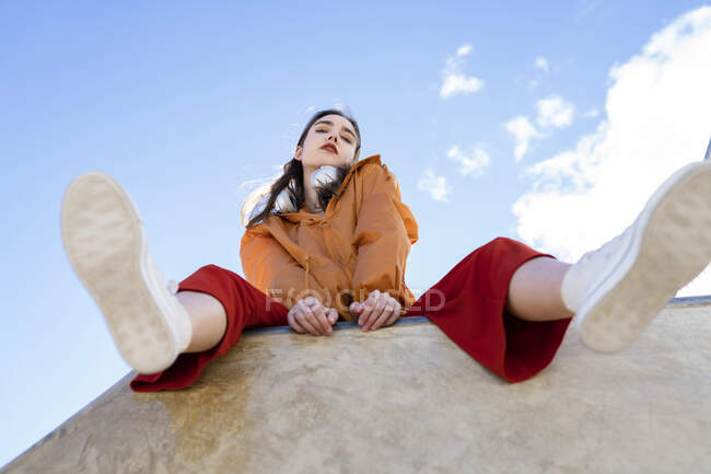 Bajo ángulo de adolescente femenino en ropa de moda y zapatos de goma mirando a la cámara desde la valla de hormigón en la espalda iluminada con cielo azul claro - foto de stock