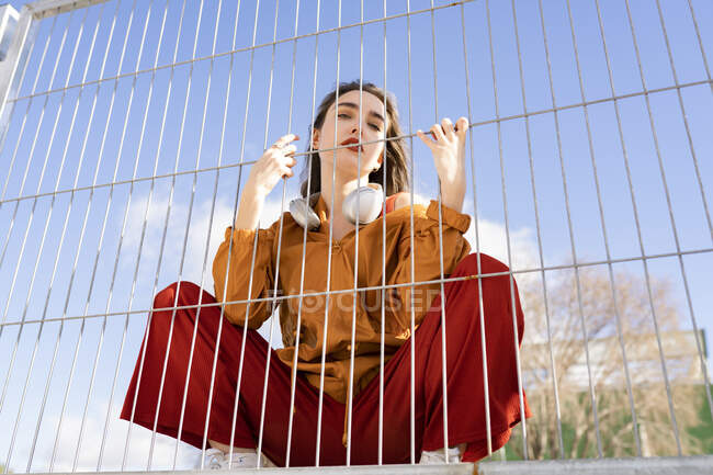 From below of cool female adolescent in stylish apparel with wireless headphones looking at camera behind metal fence in town — Photo de stock