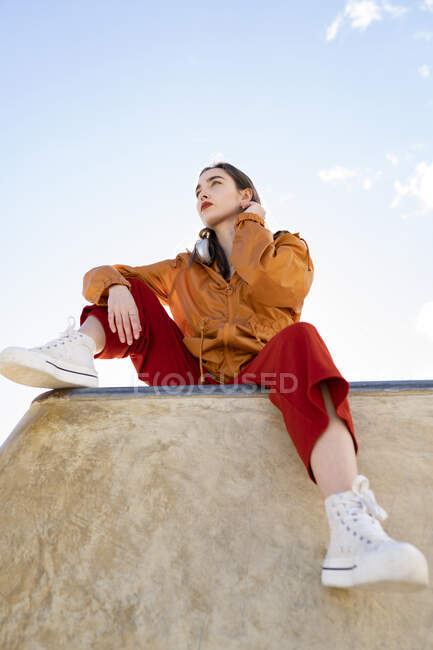 Niedriger Winkel einer Teenagerin in trendigen Klamotten und Gummischuhen, die von einem Betonzaun wegschaut, im Gegenlicht mit blauem Himmel — Stockfoto