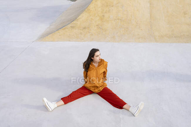 Сверху все тело молодая женщина в стильном наряде сидит в бетонном скейт-парке, глядя вдаль на солнце — стоковое фото