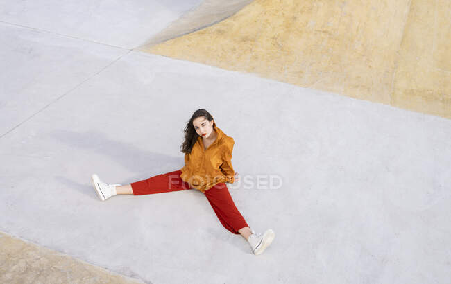 Von oben betrachtet sitzt eine junge Frau in stylischem Outfit im Betonskatepark und schaut in die Kamera im Sonnenlicht — Stockfoto