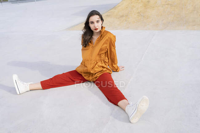 Dall'alto corpo pieno giovane donna in abito elegante seduto in skate park di cemento mentre guarda la fotocamera alla luce del sole — Foto stock