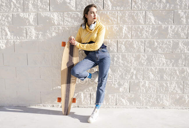 Молода модна жіноча скейтбордистка з довгим дошкою, що стоїть з піднятою ногою біля грубої стіни, дивлячись далеко — стокове фото