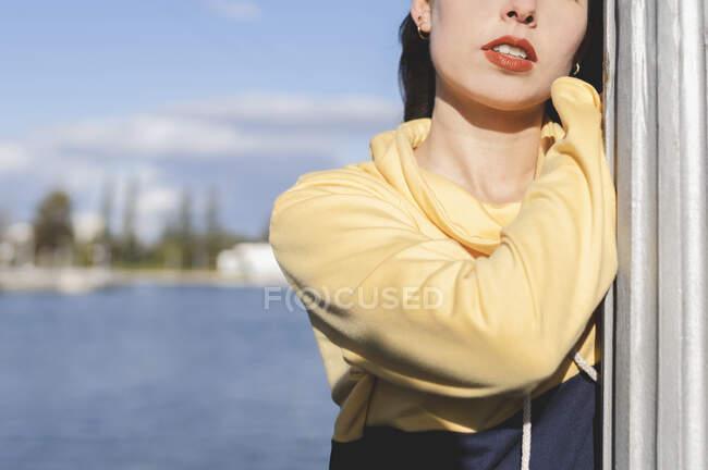 Cropped unrecognizable female adolescent in stylish outfit with red lipstick leaning on metal fence while standing against lake under blue sky — Fotografia de Stock