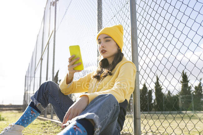 Cool female adolescent in bright casual apparel browsing internet on cellphone while leaning on grid fence in city — Photo de stock