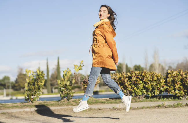 Seitenansicht einer Teenagerin in Freizeitkleidung, die in die Kamera blickt, während sie gegen den reißenden Fluss und den blauen Himmel springt — Stockfoto