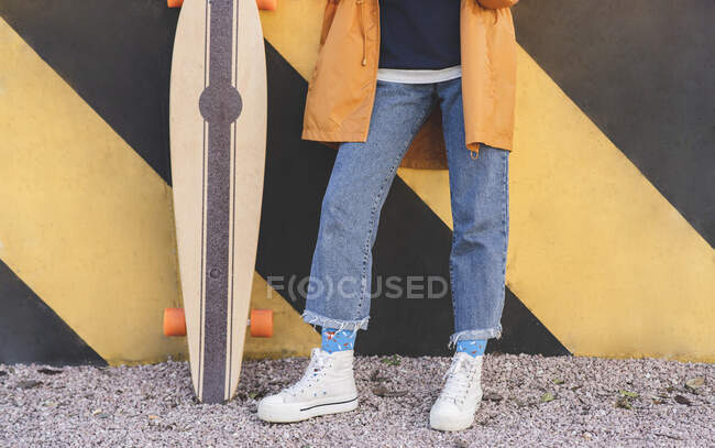 Cosecha anónima mujer con estilo en jeans de pie con longboard en la ciudad en el día soleado - foto de stock