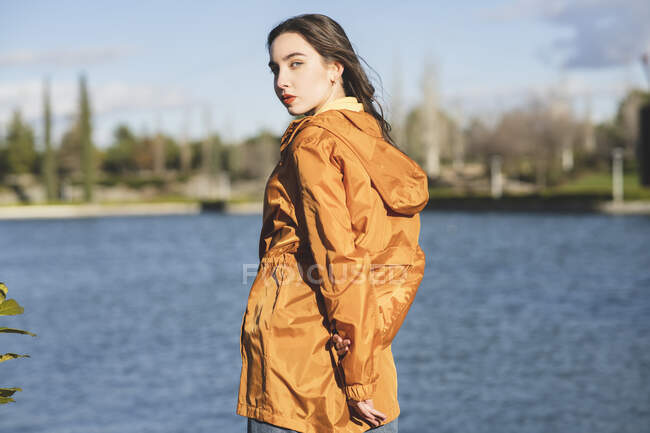 Vista posterior de la adolescente en ropa casual mirando a la cámara sobre el hombro contra el río ondulado - foto de stock