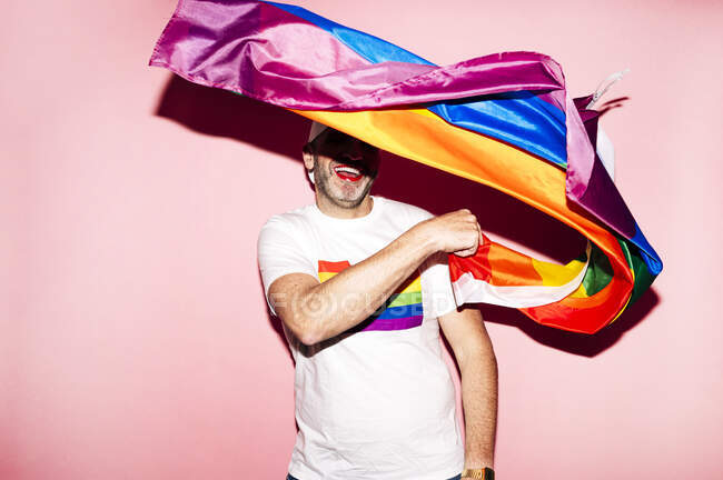 Веселый небритый гомосексуалист с красными губами в белой футболке, улыбающийся, размахивая флагом ЛГБТ на розовом фоне — стоковое фото