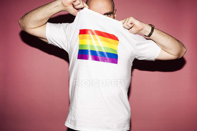 Crop anonymous young homosexual male covering face with with t shirt with rainbow flag and looking at camera — Stock Photo