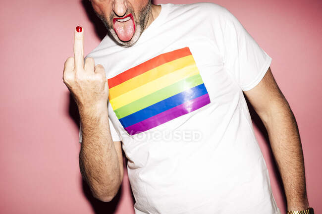Crop unrecognizable unshaven gay with red lips in with t shirt with LGBT flag showing fuck gesture with tongue out against pink background - foto de stock