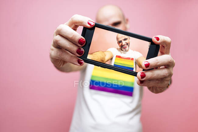 Эксцентричный взрослый лысый гей с бородой и красными губами и ногтями, одетый в белую футболку с радужным флагом и улыбающийся, делая селфи на смартфоне на розовом фоне — стоковое фото