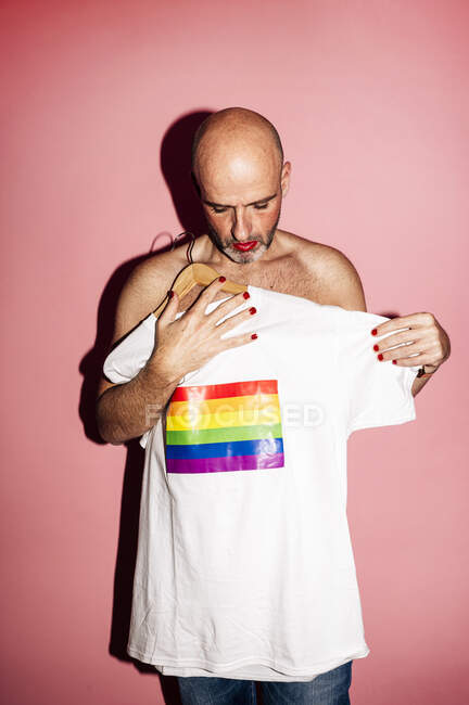 Adult bald shirtless homosexual male with red lips and nails holding white t shirt with LGBT flag against pink background — Stock Photo