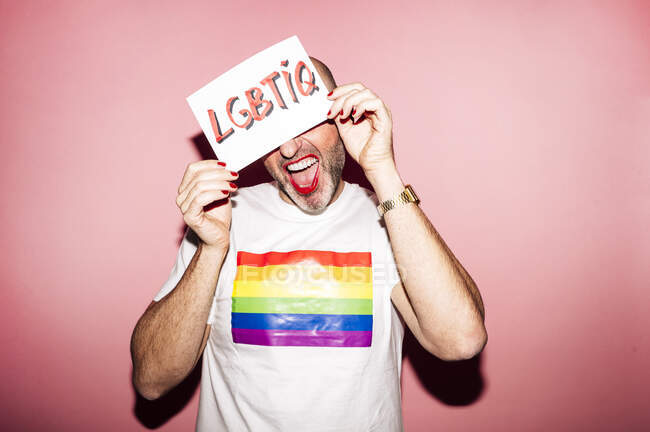 Cultivo rebelde hombre homosexual barbudo con labios rojos y manicura haciendo mueca mientras muestra y cubre la cara con papel con texto LGBTIQ sobre fondo rosa - foto de stock