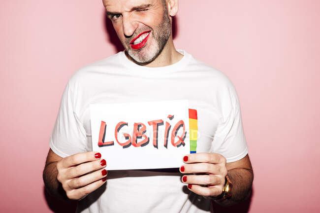 Коп бунтівний бородатий гомосексуаліст з червоними губами і манікюр робить сірники, показуючи папір з LGBTIQ текст на рожевому фоні — стокове фото