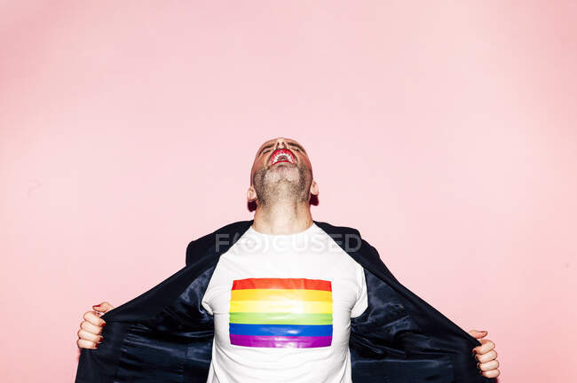 Confident bearded man with red lips screaming and demonstrating LGBT flag on white t shirt against pink background — Stock Photo