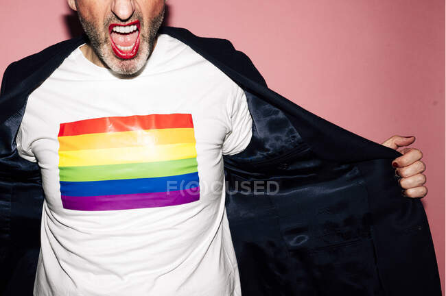 Gestutzter, bis zur Unkenntlichkeit selbstbewusster bärtiger Mann mit roten Lippen, der schreit und LGBT-Flagge auf weißem T-Shirt vor rosa Hintergrund zeigt — Stockfoto