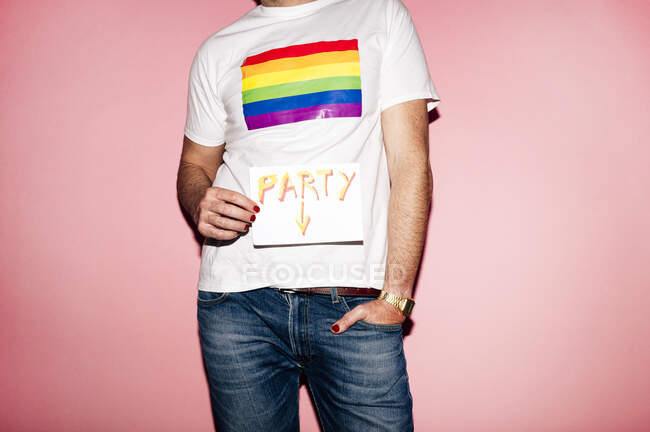 Crop unrecognizable homosexual guy in white t shirt with rainbow flag standing against pink background and showing paper with Party inscription — Fotografia de Stock