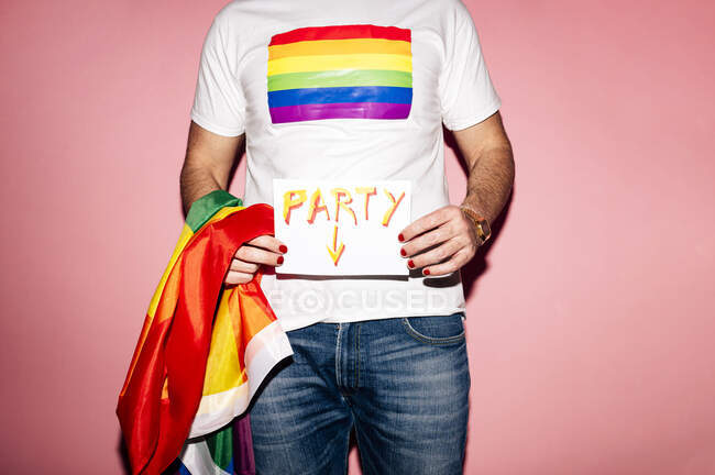 Crop unrecognizable homosexual guy in white t shirt with rainbow flag standing against pink background and showing paper with Party inscription — Photo de stock