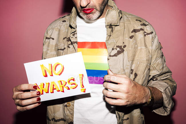 Crop anonymous bearded eccentric middle aged man in shirt with LGBT flag with makeup and manicure showing paper with No Wars inscription against pink background — Photo de stock