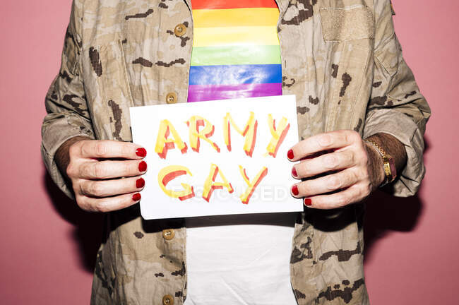 Анонимный бородатый эксцентричный мужчина средних лет в футболке с флагом ЛГБТ с маникюром, показывающим бумагу с армейскими надписями геев на розовом фоне — стоковое фото