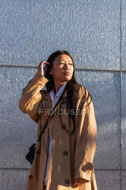Serious Asian female in stylish outfit with long hair looking away while standing near modern building — Stock Photo