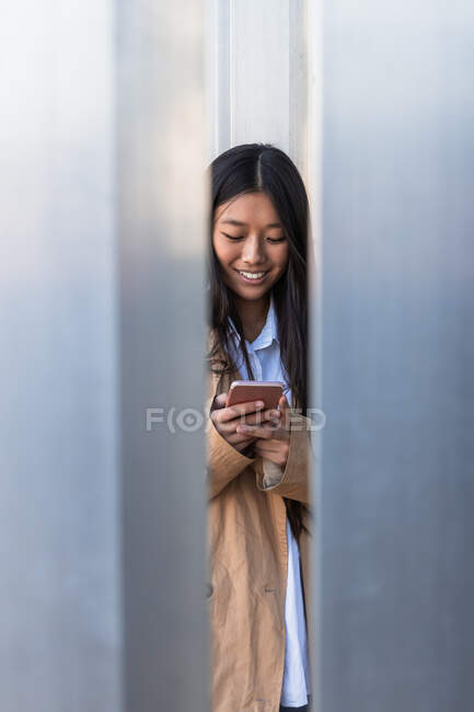 Full length of happy Asian female in stylish outfit standing on street and browsing internet on mobile phone — Fotografia de Stock