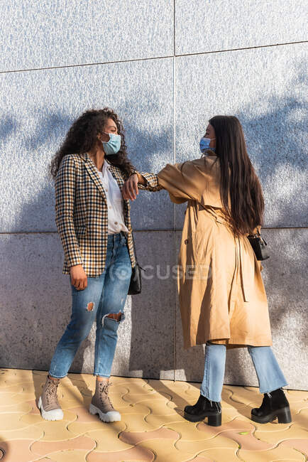 Вид збоку на етнічних друзів у повсякденному одязі з медичними масками, які дивляться один на одного під час привітання з ліктями — стокове фото