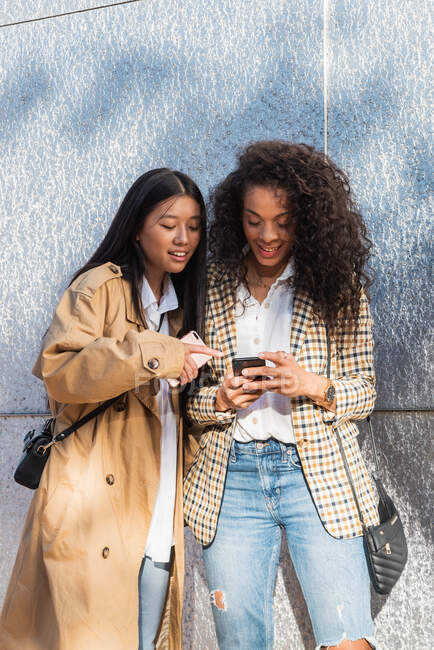 Optimistic ethnic female friends in trendy outfits standing on city street and chatting on mobile phone while having break — Photo de stock
