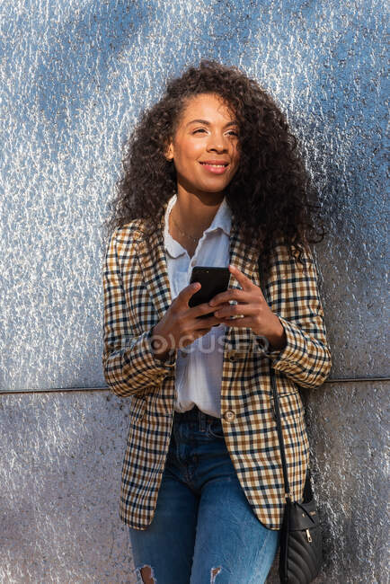 Mujer afroamericana en traje de moda con pelo rizado teléfono de navegación mientras está de pie en la calle cerca de la pared de hormigón - foto de stock