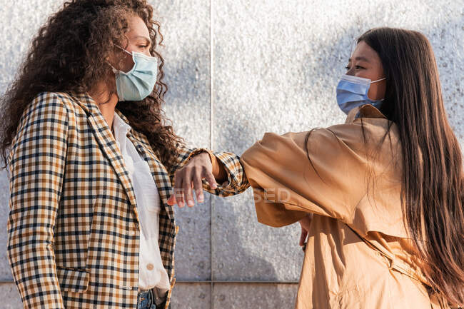Вид сбоку на друзей-этнических женщин в повседневной одежде в медицинских масках, смотрящих друг на друга во время приветствия локтями — стоковое фото