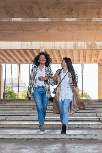 Delighted female friends with toothy smiles wearing trendy clothes looking away while walking together stairway in town — Stock Photo