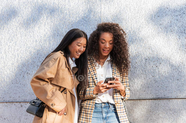 Optimistic ethnic female friends in trendy outfits standing on city street and chatting on mobile phone while having break — Stock Photo