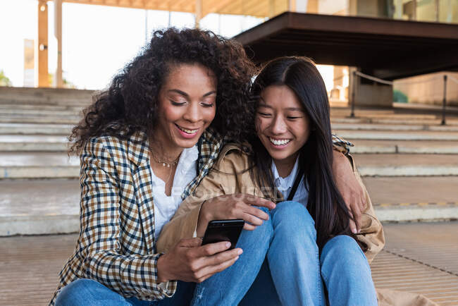 Optimistische ethnische Freundinnen in trendigen Outfits sitzen auf der Straße und chatten auf dem Handy — Stockfoto