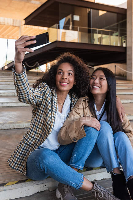 Optimistic ethnic female friends in trendy outfits sitting on city street and taking selfie while having break — Photo de stock