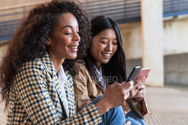 Optimistische ethnische Freundinnen in trendigen Outfits sitzen auf der Straße der Stadt und chatten während der Pause mit dem Handy — Stockfoto