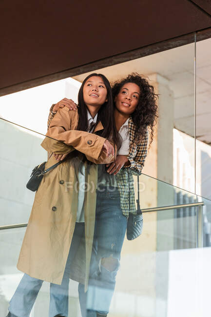 Pleasant ethnic female friends in stylish clothes with toothy smiles looking away while leaning on glass railing — Fotografia de Stock