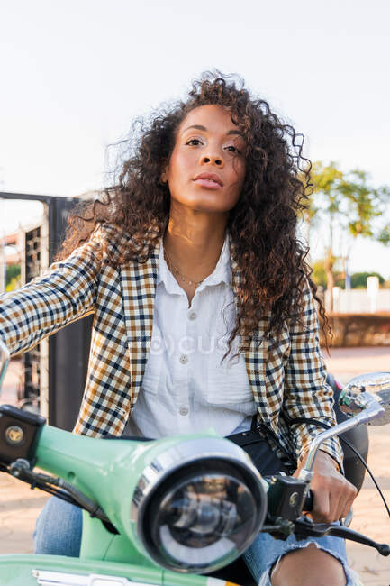 Young wistful black female in trendy outfit looking away while riding scooter on sunny day — Photo de stock