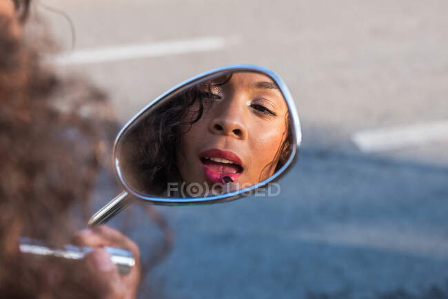 Dreamy African American female with curly hair looking in side mirror of scooter and applying pink lipstick — Photo de stock