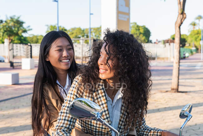 Optimistic Asian female with toothy smile and satisfied African American woman with curly hair looking at each other while sitting on scooter — Photo de stock