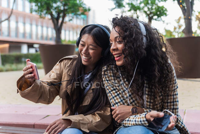 Asiática hembra divertirse llamada en el teléfono inteligente y centrado negro mujer escuchando música y navegar por Internet en el teléfono móvil - foto de stock